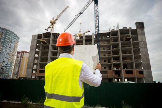 Rear view photo of male construction engineer with blueprints controlling work on building site